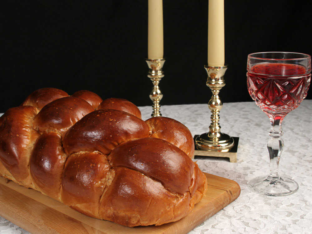 Baked challah bread with cover, two Shabbat candles and Kiddush cup are set upon a dinner table.